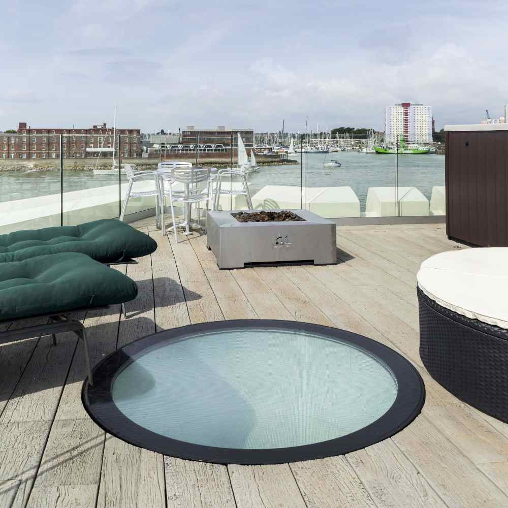 A rooftop terrace features a glass skylight, lounge chairs, a round ottoman, a fire pit, and a bar table with stools. The background shows a harbor with boats and buildings under a cloudy sky.