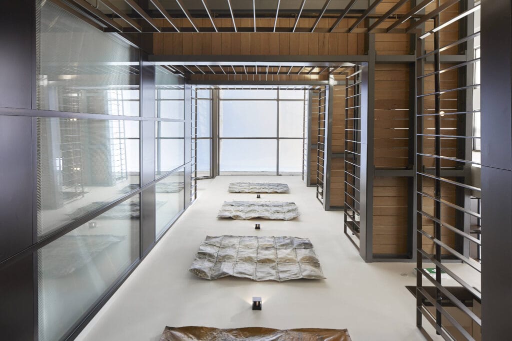 A modern minimalist room features three large abstract canvases on the floor, flanked by metallic and glass walls with a grid-like ceiling structure.