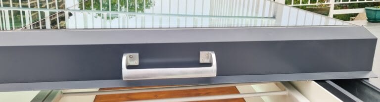 A close-up view of a metal handle on a railing structure with glass panels and greenery in the background.