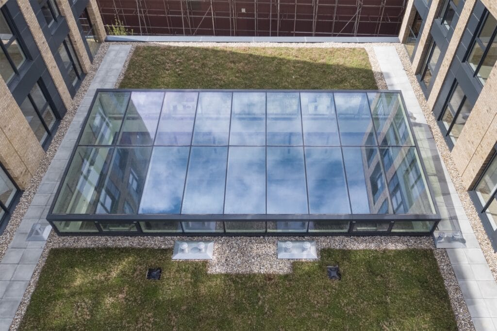 Aerial view of a glass skylight roof on a modern building, surrounded by a grassy courtyard and stone pathways.