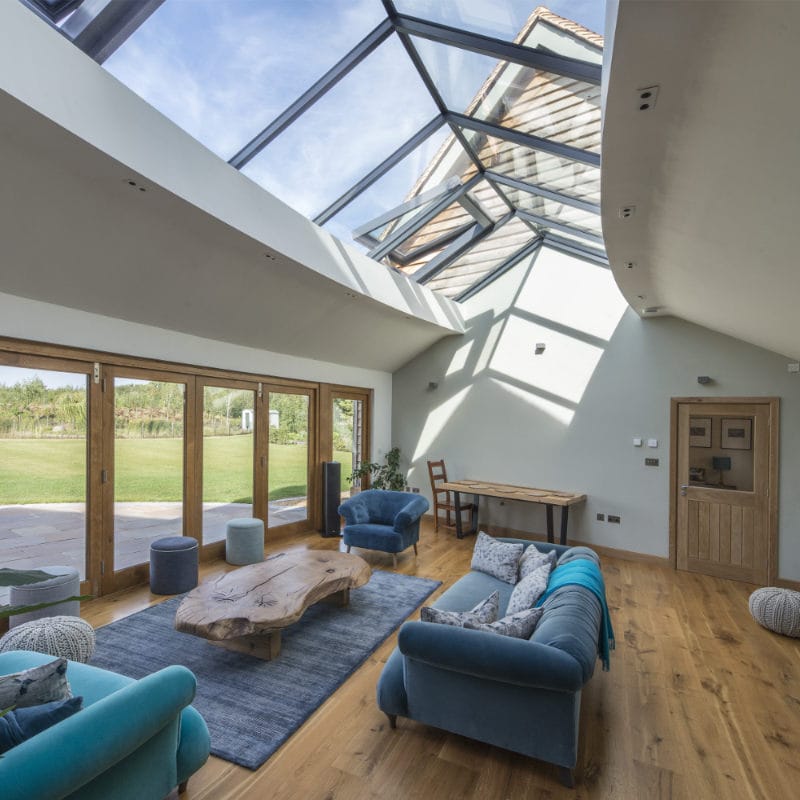IW-130916-VIS-079 A spacious living room with a glass ceiling, wooden floors, blue and grey furniture, a wooden coffee table, and a dining table with chairs. Large windows allow natural light to illuminate the room.