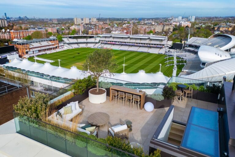 Rooftop terrace with seating and plants overlooks a large cricket stadium and cityscape in the background.