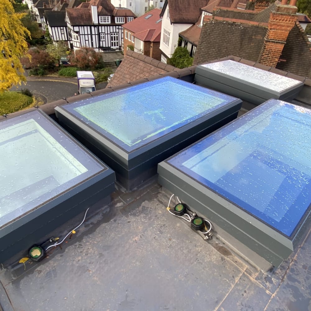 NGR Electric Opening Rooflights With Sensors Rooftop view showing several rectangular skylights with rain droplets on them, surrounded by a residential neighborhood with houses and trees.