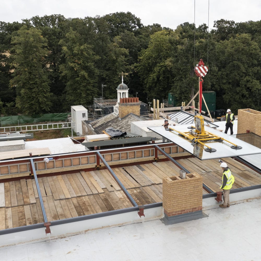 NGR High End Residential Two Ton Elevated Rooflight installation via Crane Construction workers install large glass panels using a crane on a rooftop, surrounded by greenery and scaffolding on nearby structures.