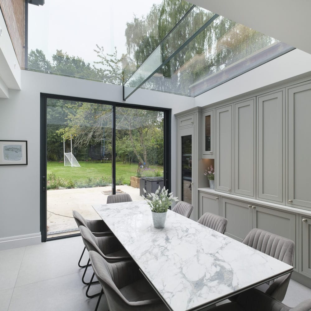 NGR Multi Part Rooflight with Glass Rafters High End Commercial A modern kitchen and dining area with a large glass skylight, marble dining table, and sliding glass doors leading to a garden with a soccer goal and trees in the background.