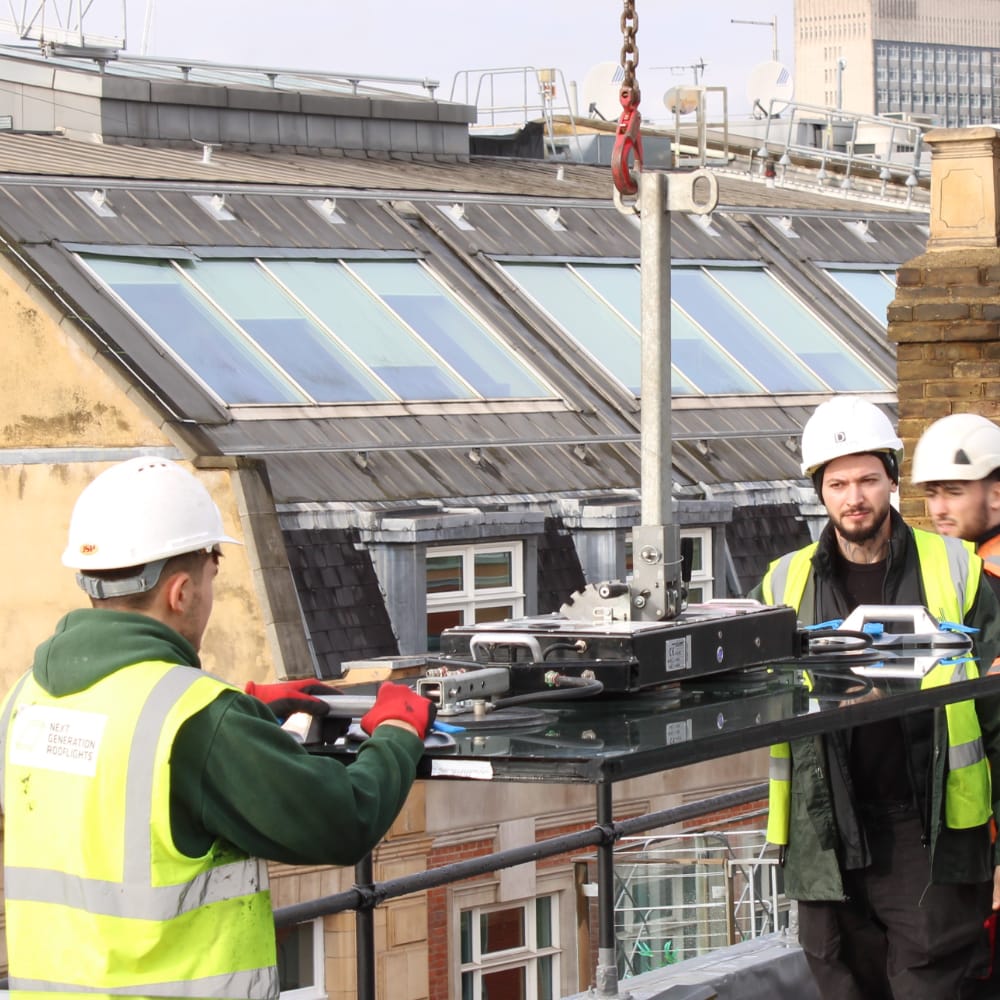 NGR Rooflight being installed Three construction workers wearing safety gear are installing a large glass panel on the exterior of a building using a hoisting mechanism. Rooftop and windows of nearby buildings are visible in the background.