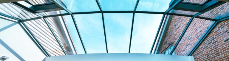 Glass ceiling structure with black metal frames, providing a view of a clear blue sky. The surrounding building walls are made of brick and metal.