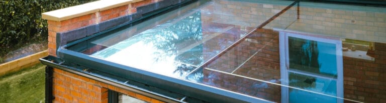 A close-up view of a modern building extension with a glass roof, showing reflections on the glass and brick walls. Greenery is visible in the background.