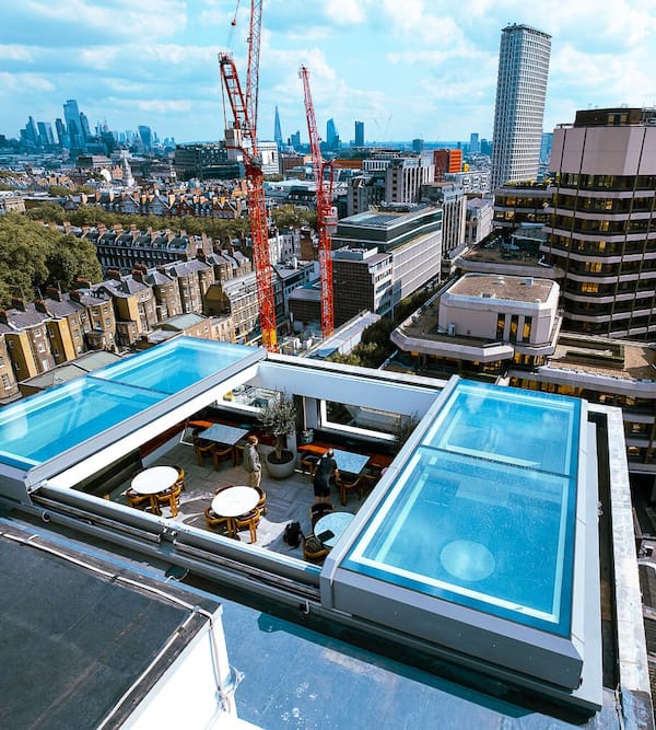 ventilation Rooftop view of a city with a transparent glass pool and seating area, surrounded by buildings and cranes, under a partly cloudy sky.